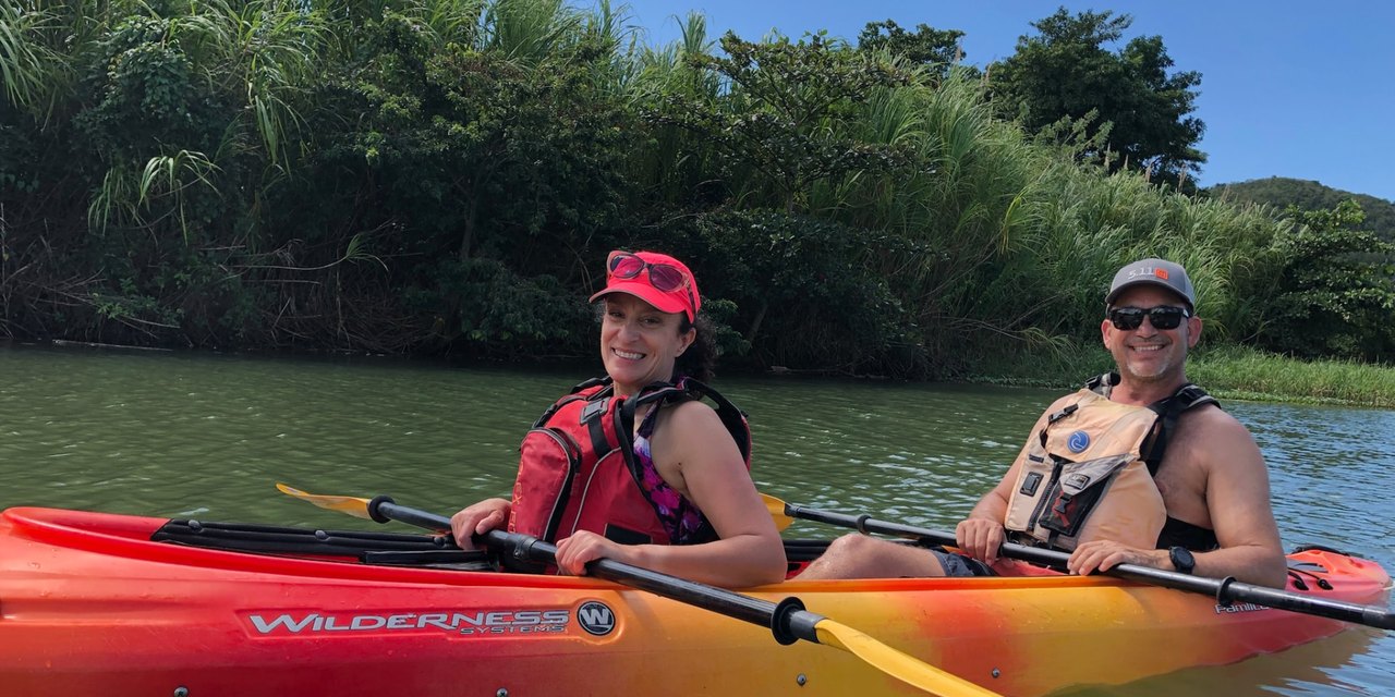 Todd and Deanna Valdez smiling together in a two-person kayak on calm green water, paddles in hand, with a dense green treeline behind them.
