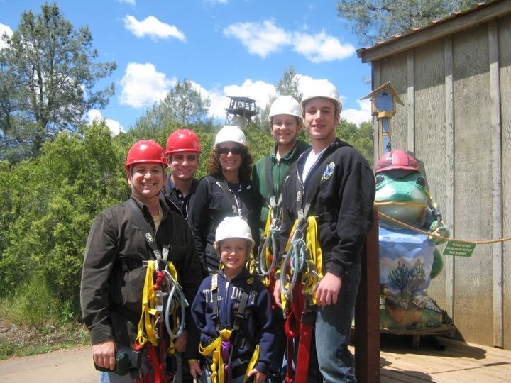 Todd Valdez with a group in helmets and climbing harnesses at an outdoor course among pine trees.