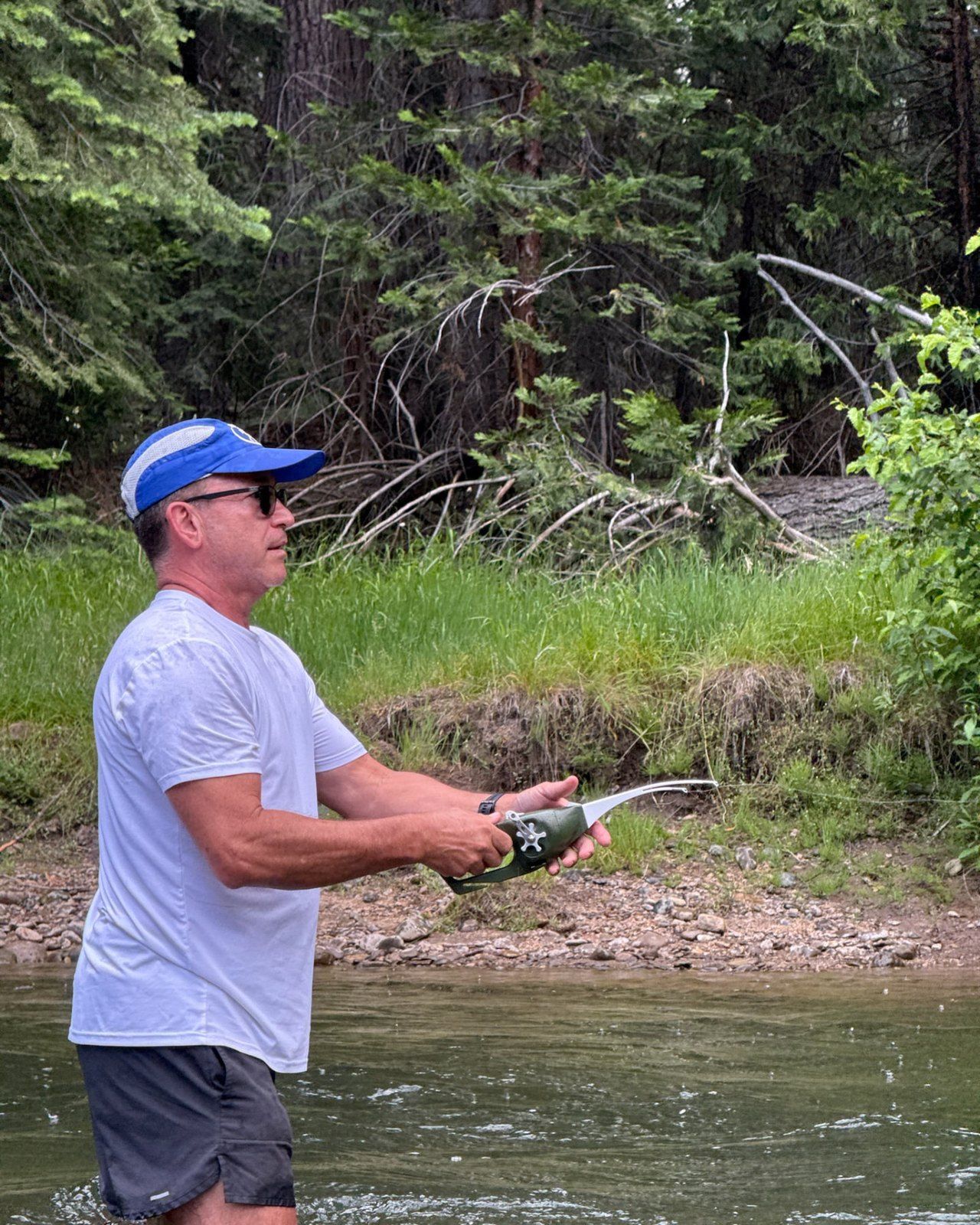 A person standing in shallow river water, fishing with a rod, with a green wooded bank behind.