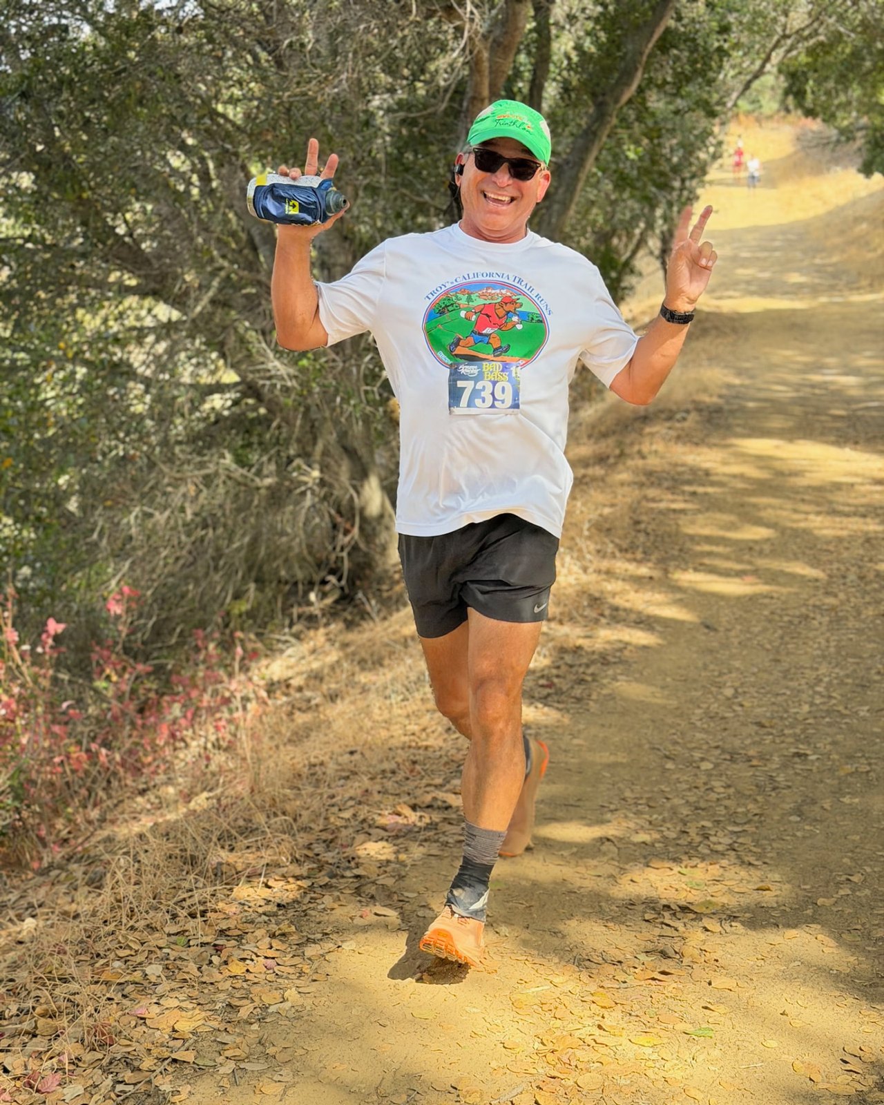 Todd Valdez running on a shaded dirt trail in a race shirt and cap.